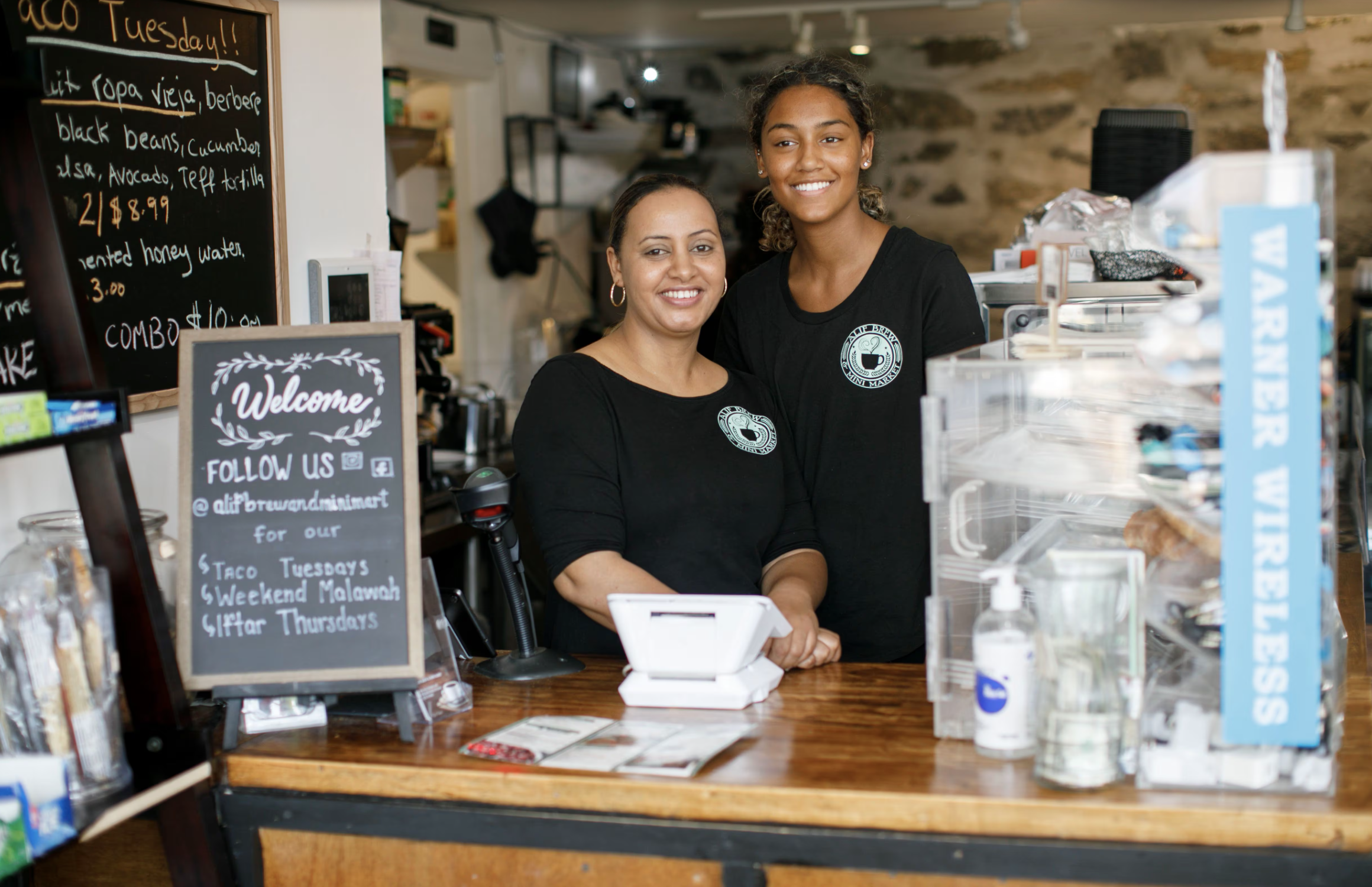 Shop owner Hayat Alif (left) and her daughter Aida Solomonat (right) stand behind the counter at their Ethiopian coffee shop, smiling at the camera